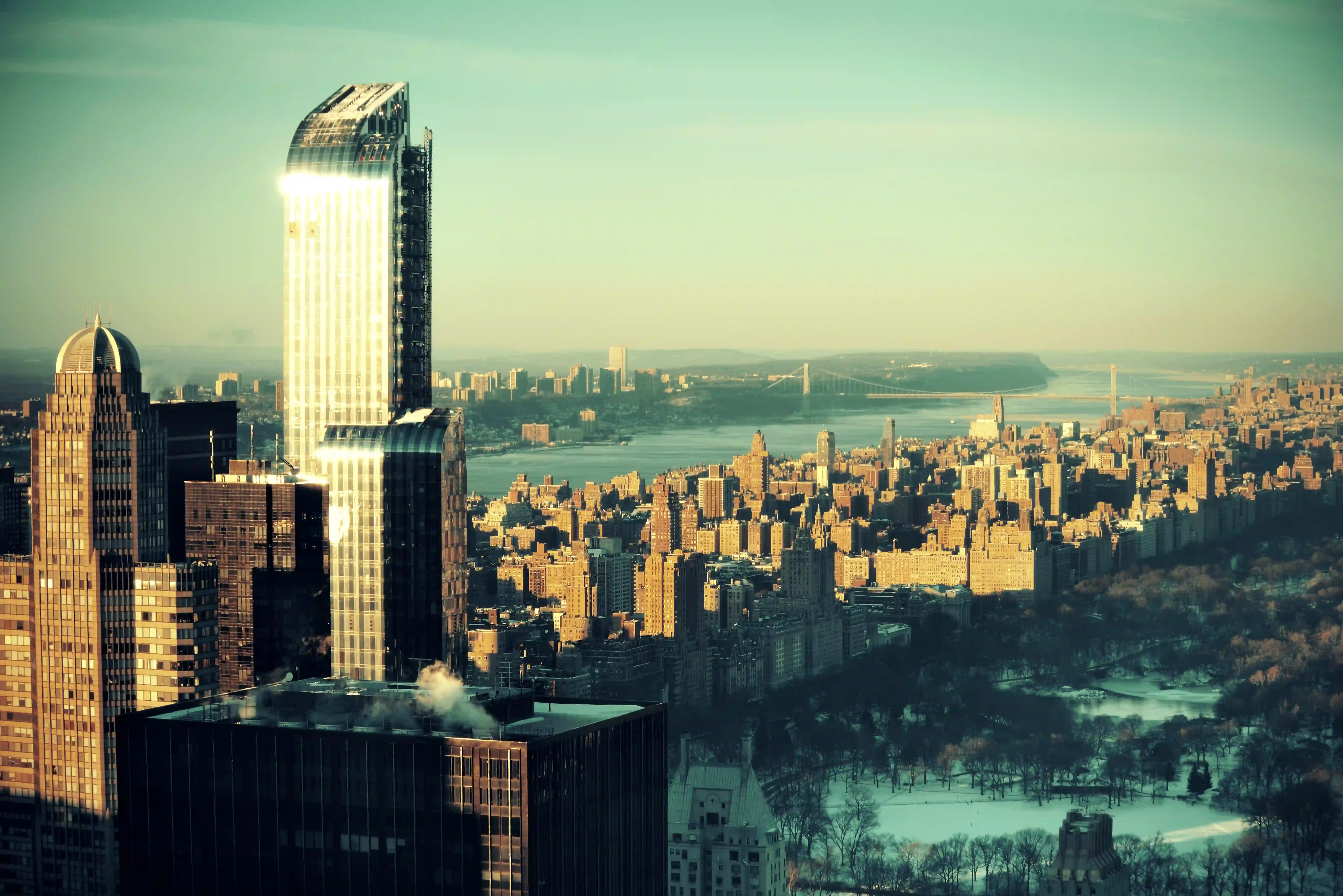 Aerial view of New York City skyline with sunlit skyscrapers, Central Park in winter, and distant river with bridges.
