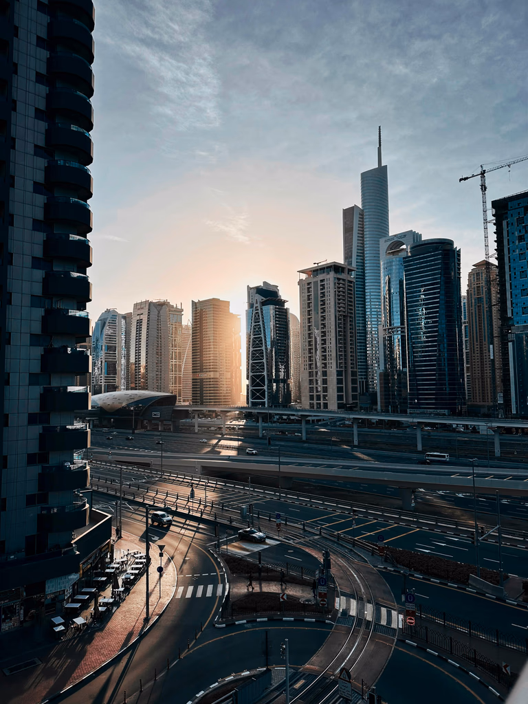 Cityscape at sunrise with modern skyscrapers, highways, and curved roads casting long shadows.