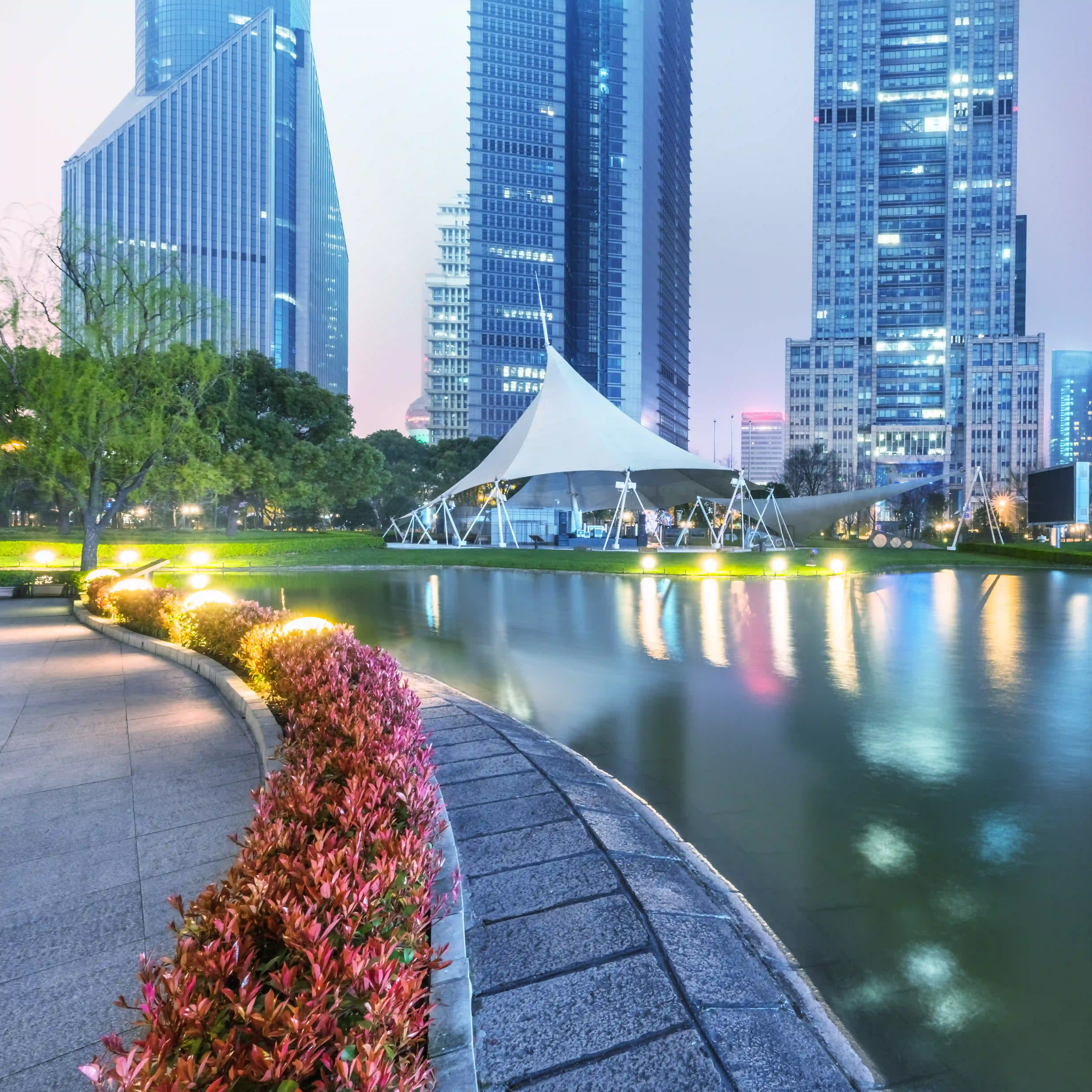 Urban park at dusk with illuminated pathway, colorful bushes, water reflecting city skyscrapers, and a large white event tent.