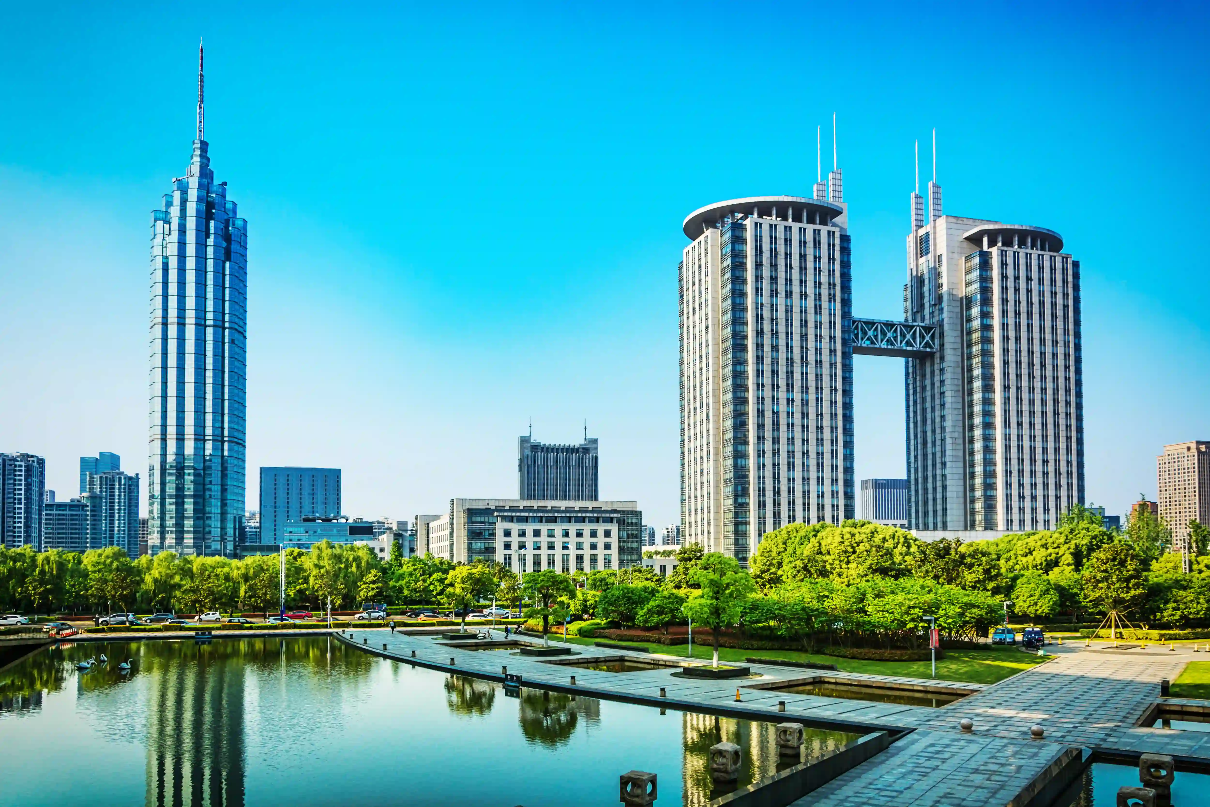 Modern cityscape with tall skyscrapers, a reflecting pond, and green trees under clear blue sky.