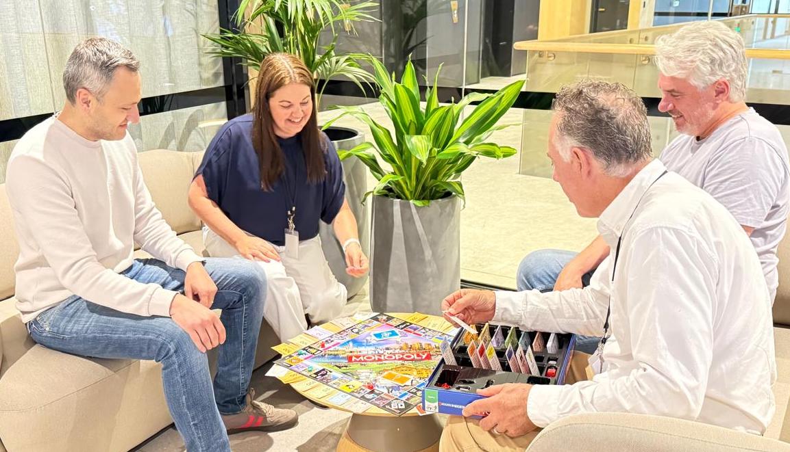 Four adults sitting around a table playing a Monopoly board game indoors with a potted plant in the background.