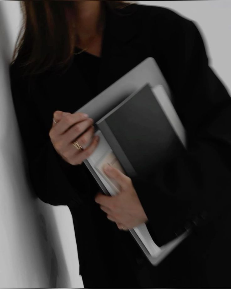 Person in black outfit holding a laptop, a black notebook, and some papers against a white wall.