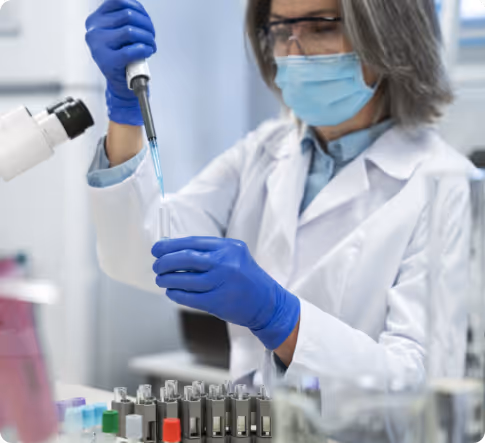 Scientist in a lab coat, gloves, mask, and safety glasses using a pipette to transfer liquid into a test tube in a laboratory.
