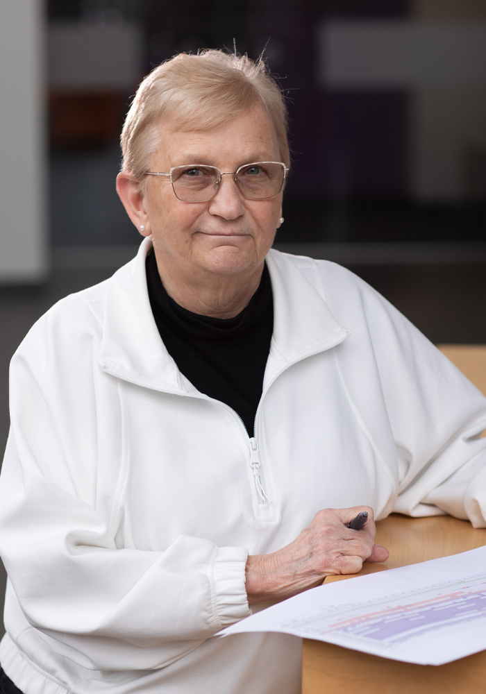 Elderly woman wearing glasses and a white jacket sitting at a table holding a pen and a document.