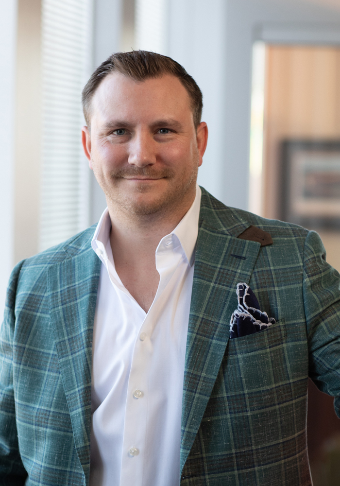 Smiling man wearing a green plaid blazer and white dress shirt standing indoors near a window.