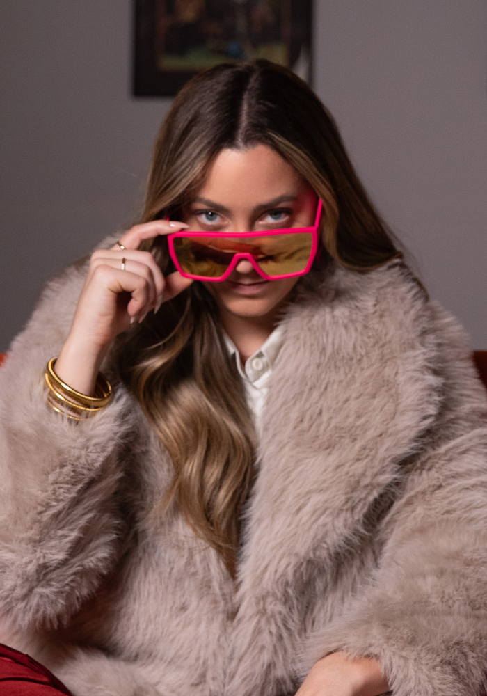 Woman with long hair wearing a fluffy beige coat and pink-tinted sunglasses, looking over the top of her glasses.
