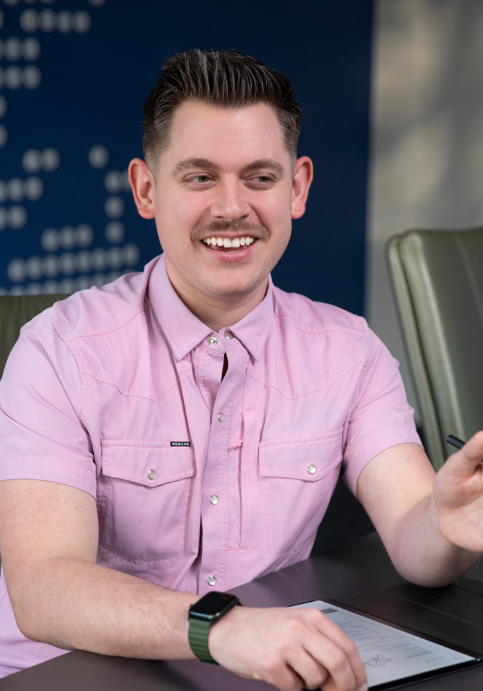 Smiling man with mustache wearing a pink button-up shirt and a smartwatch seated at a table with a tablet.