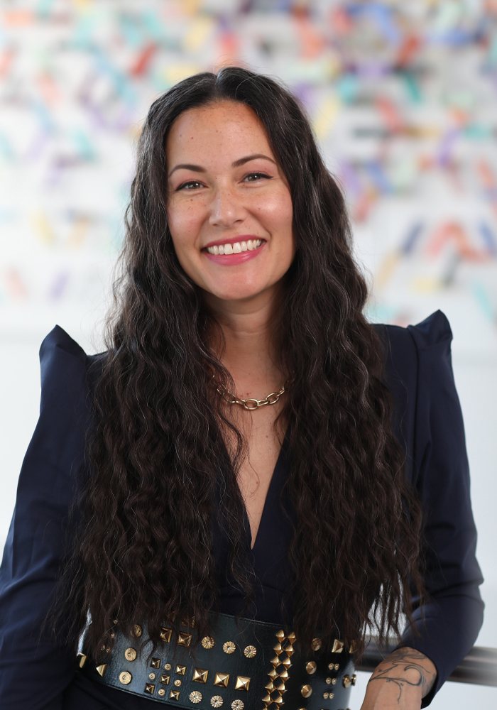 Smiling woman with long curly dark hair wearing a navy blue blouse and studded black belt against a colorful blurred background.