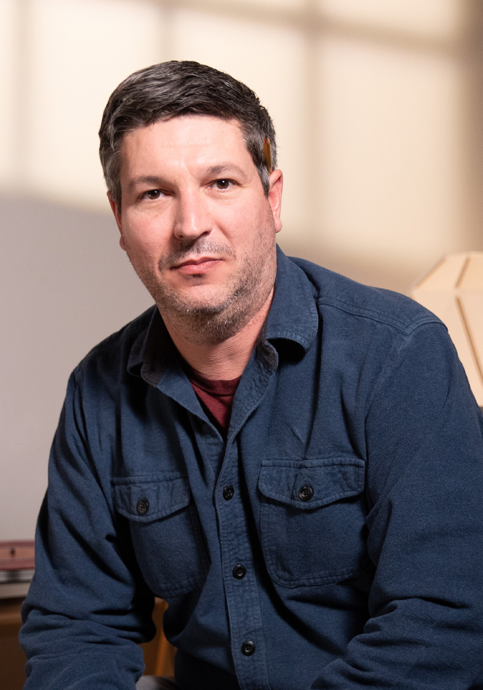 Man with short dark hair and light beard wearing a dark blue button-up shirt sitting indoors.