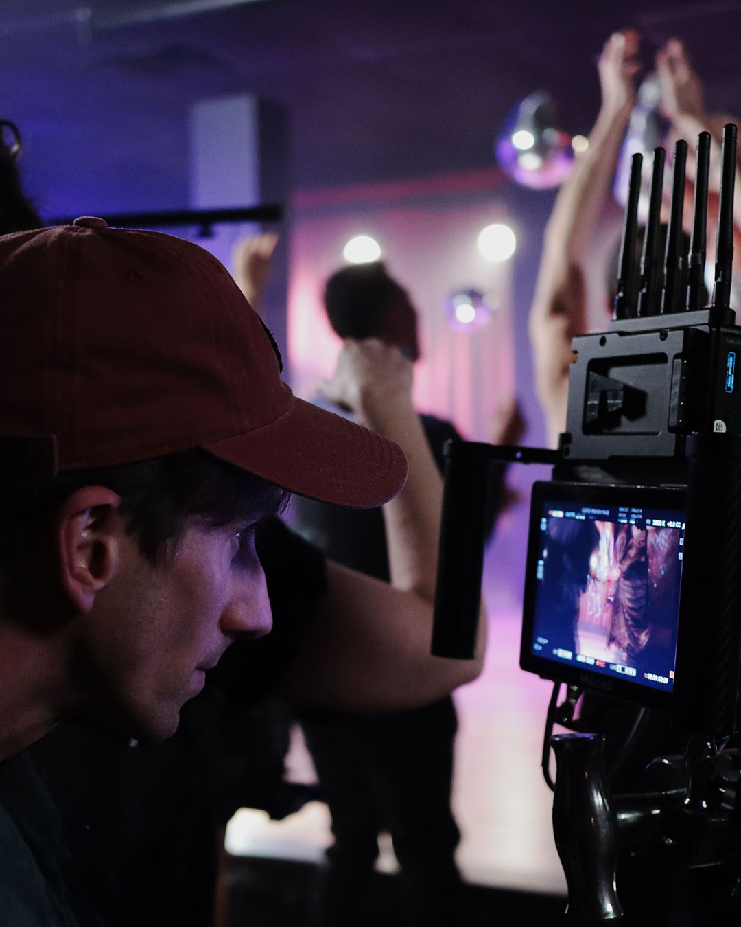 Man wearing red cap closely monitoring a video camera screen during a dimly lit event or production.