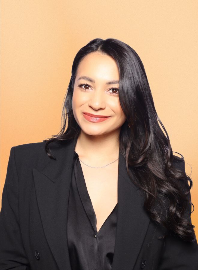 Smiling woman with long dark hair wearing a black blazer and blouse against a peach background.