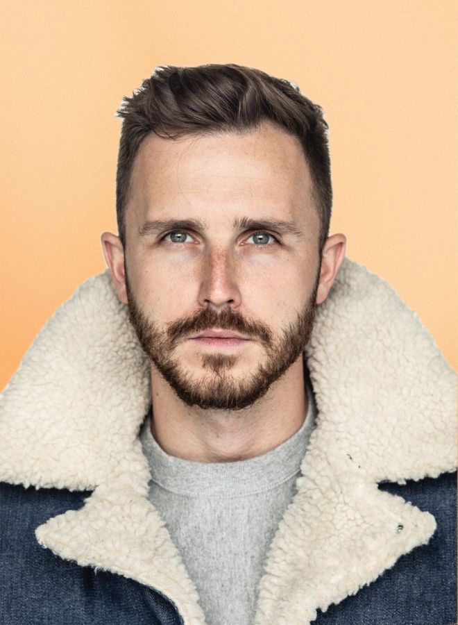 Close-up portrait of a man with short brown hair and beard wearing a fluffy lined denim jacket against a light orange background.
