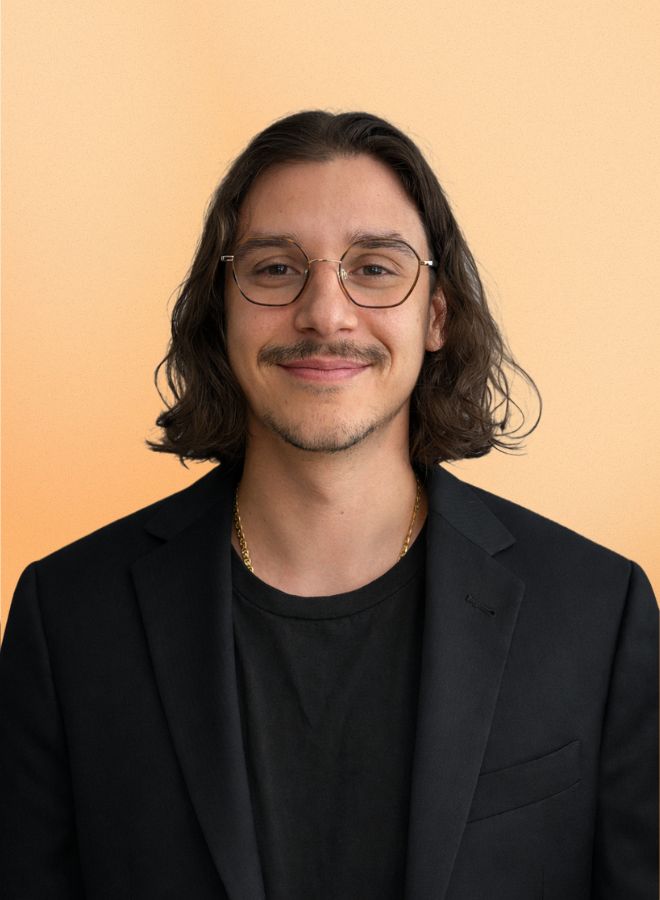 Man with shoulder-length dark hair and glasses wearing a black blazer and t-shirt, smiling against a peach background.