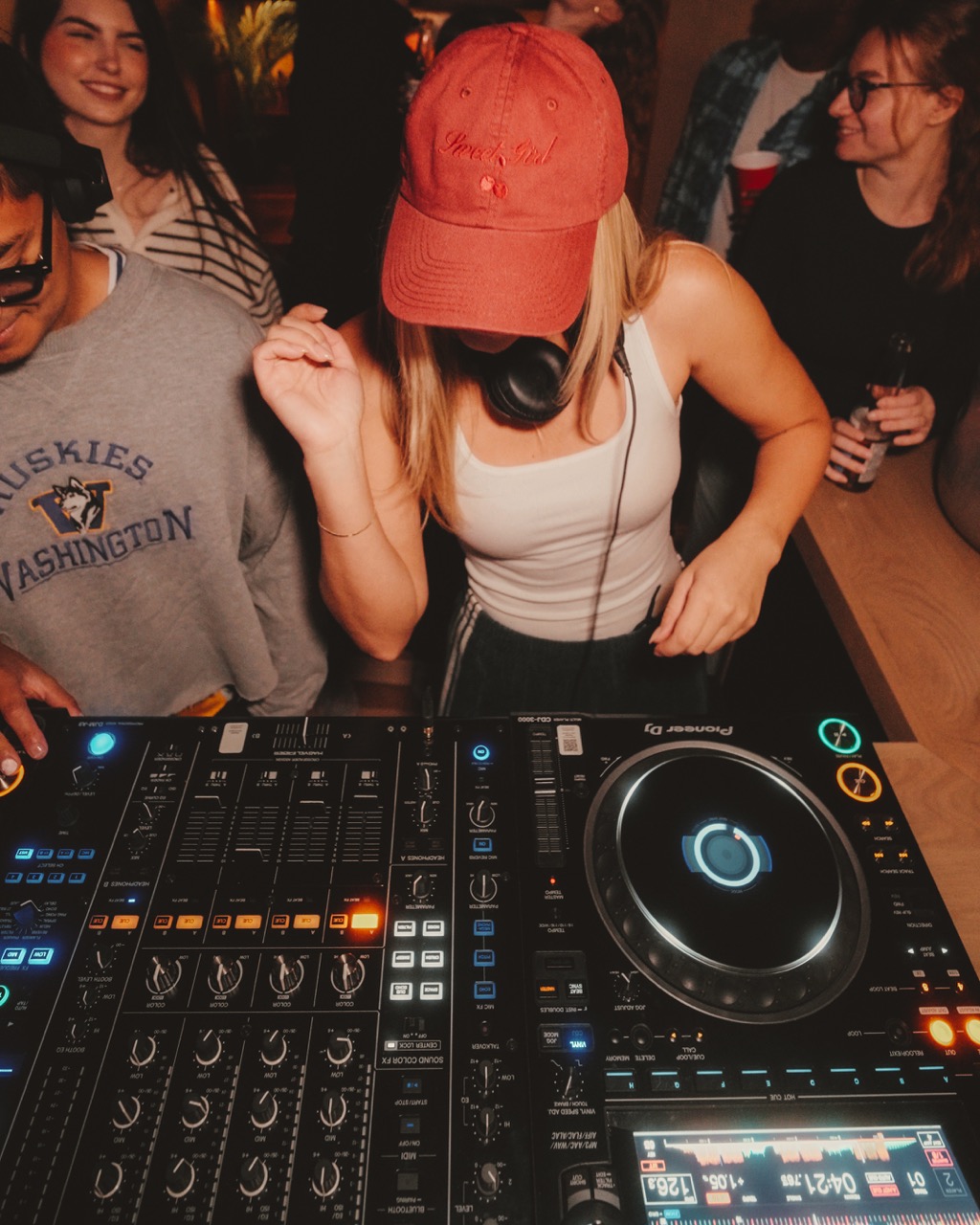 DJ wearing a red cap and white tank top mixing music on a Pioneer DJ controller at a party with people dancing nearby.