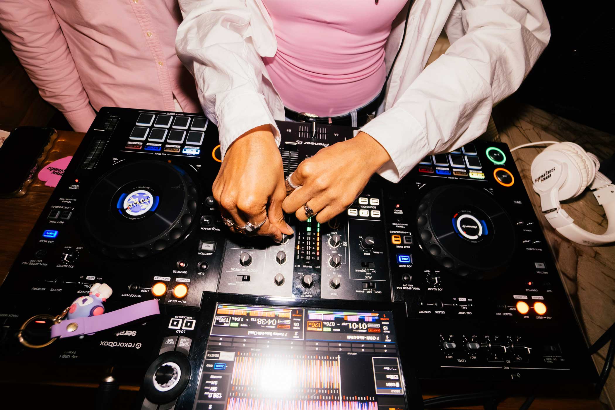 Person adjusting controls on a Pioneer DJ mixer with white headphones and a pink keychain nearby.