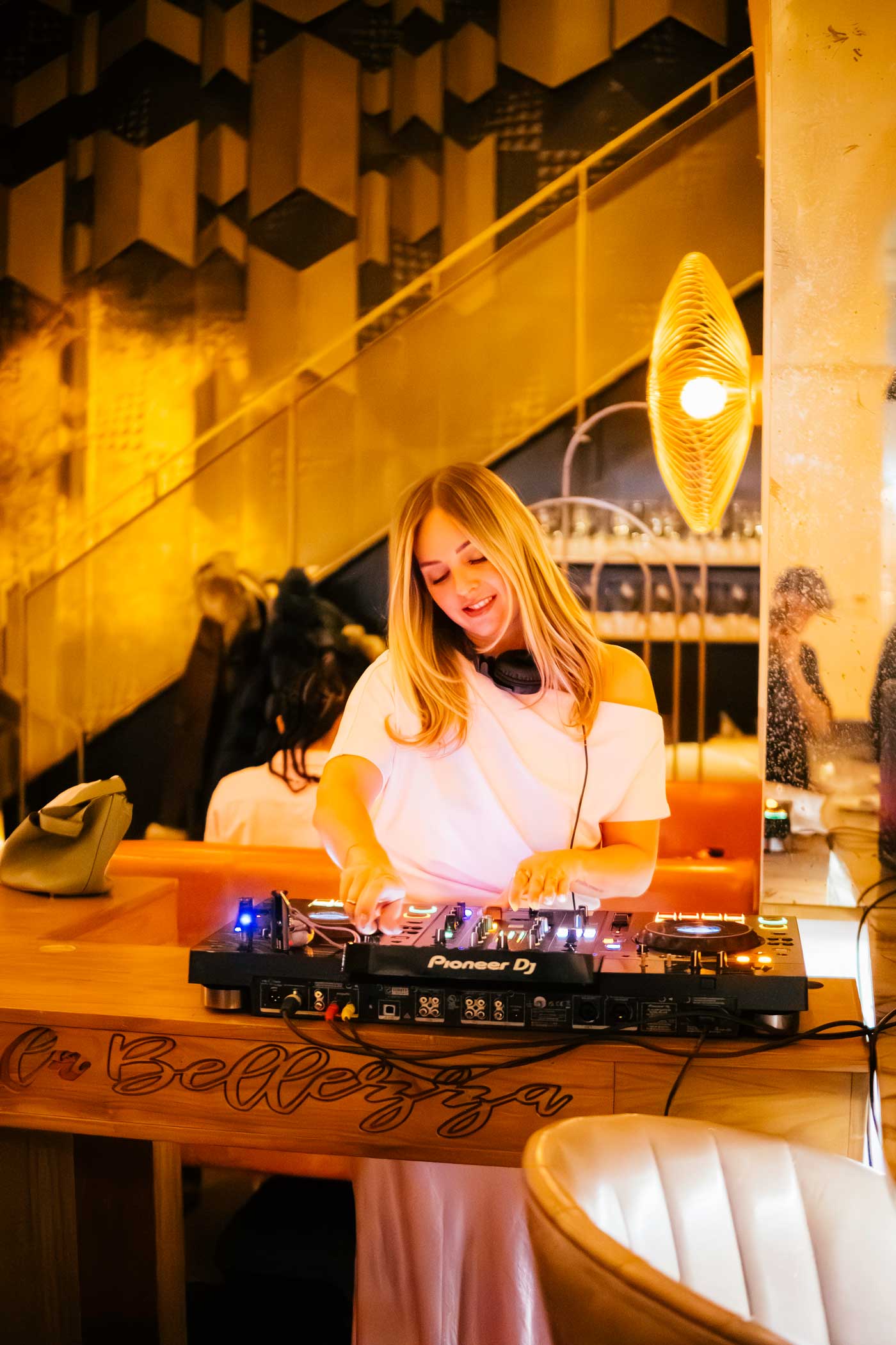 Smiling female DJ in white dress playing music on a Pioneer DJ controller in a warmly lit modern venue.