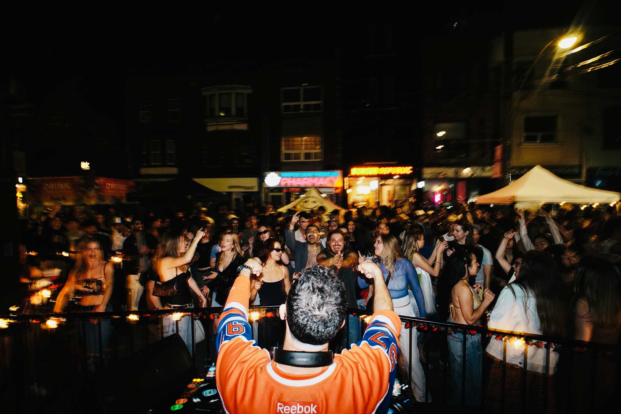 DJ in an orange Reebok jersey performing for a lively crowd dancing outdoors at night.