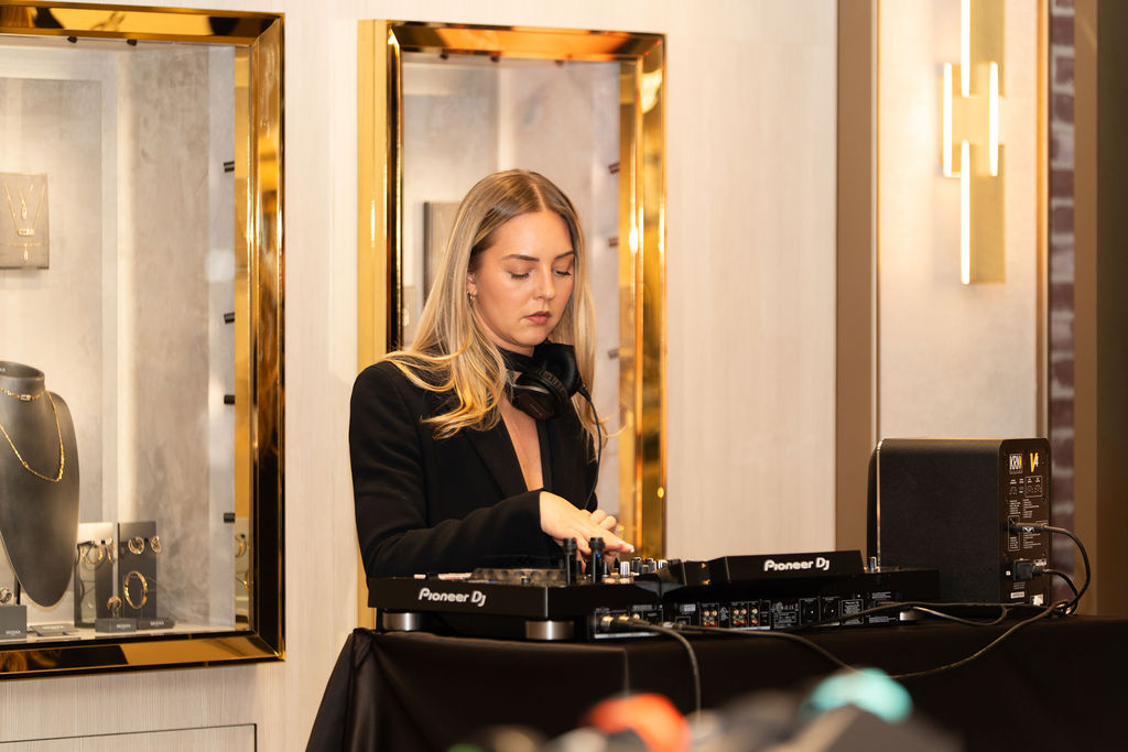 A female DJ with headphones around her neck mixing music on a Pioneer DJ controller in a stylish room with gold-framed display cases and modern lighting.