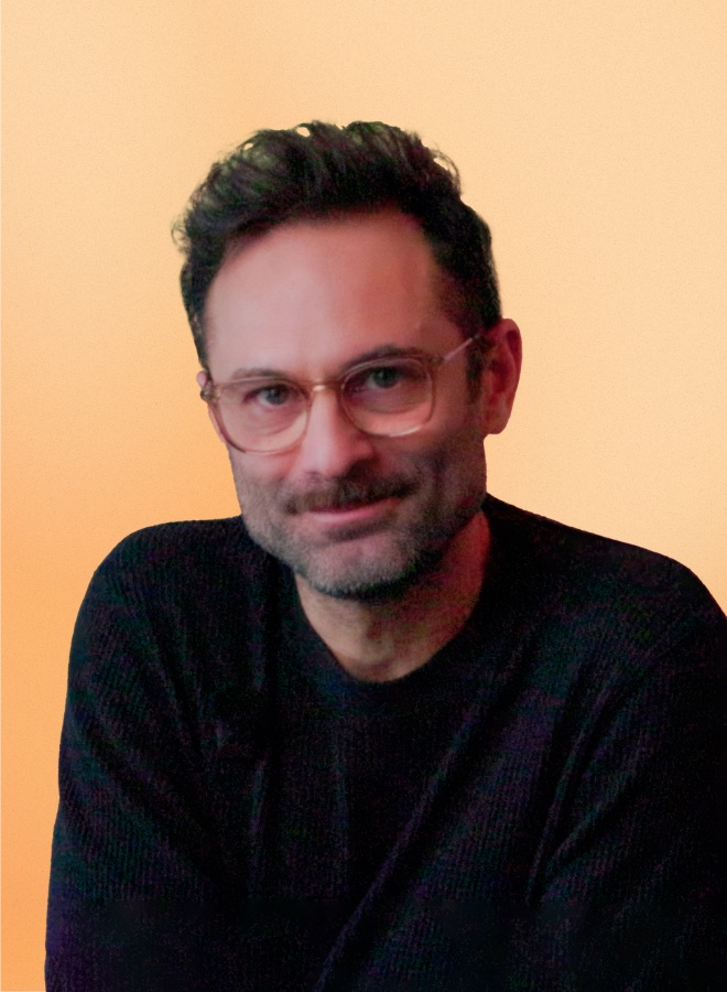 Man with short dark hair, wearing glasses and a black sweater, smiling against a light orange background.