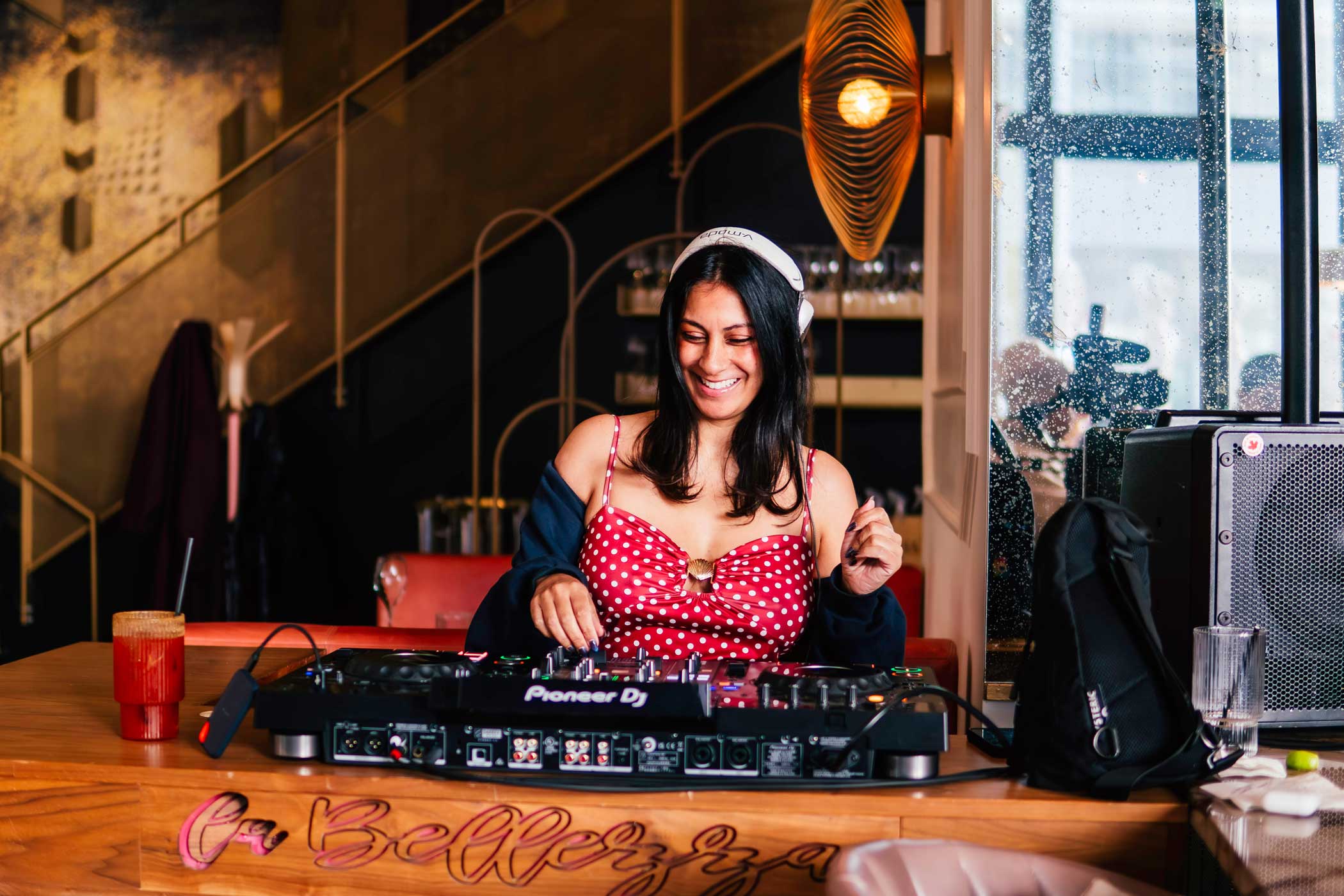 Smiling woman DJ with headphones mixing music on Pioneer DJ controller at a wooden bar table.
