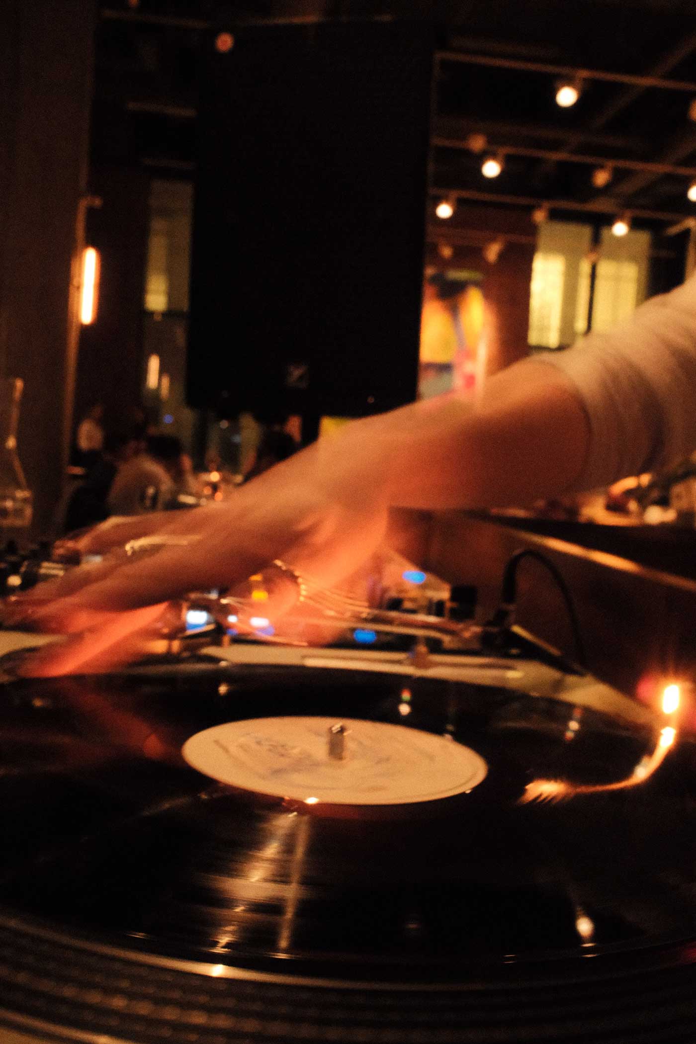 DJ's hand adjusting a vinyl record on a turntable in a dimly lit bar with blurred background of people and lights.