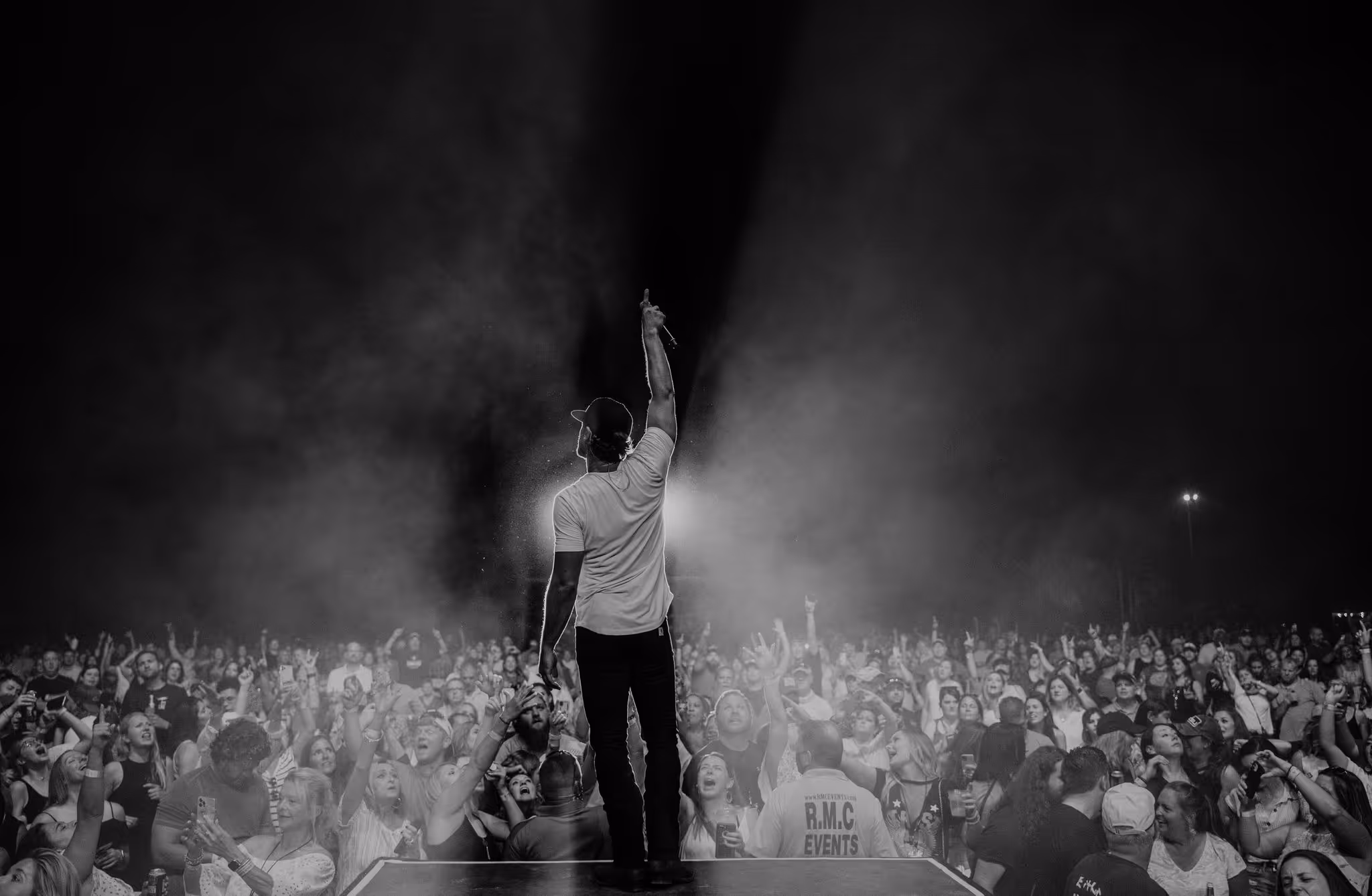 An artist stands on a stage with his back to the camera holding his hand in the air with his finger pointing up while being silhouetted by a stage light  in front a cheering audience.