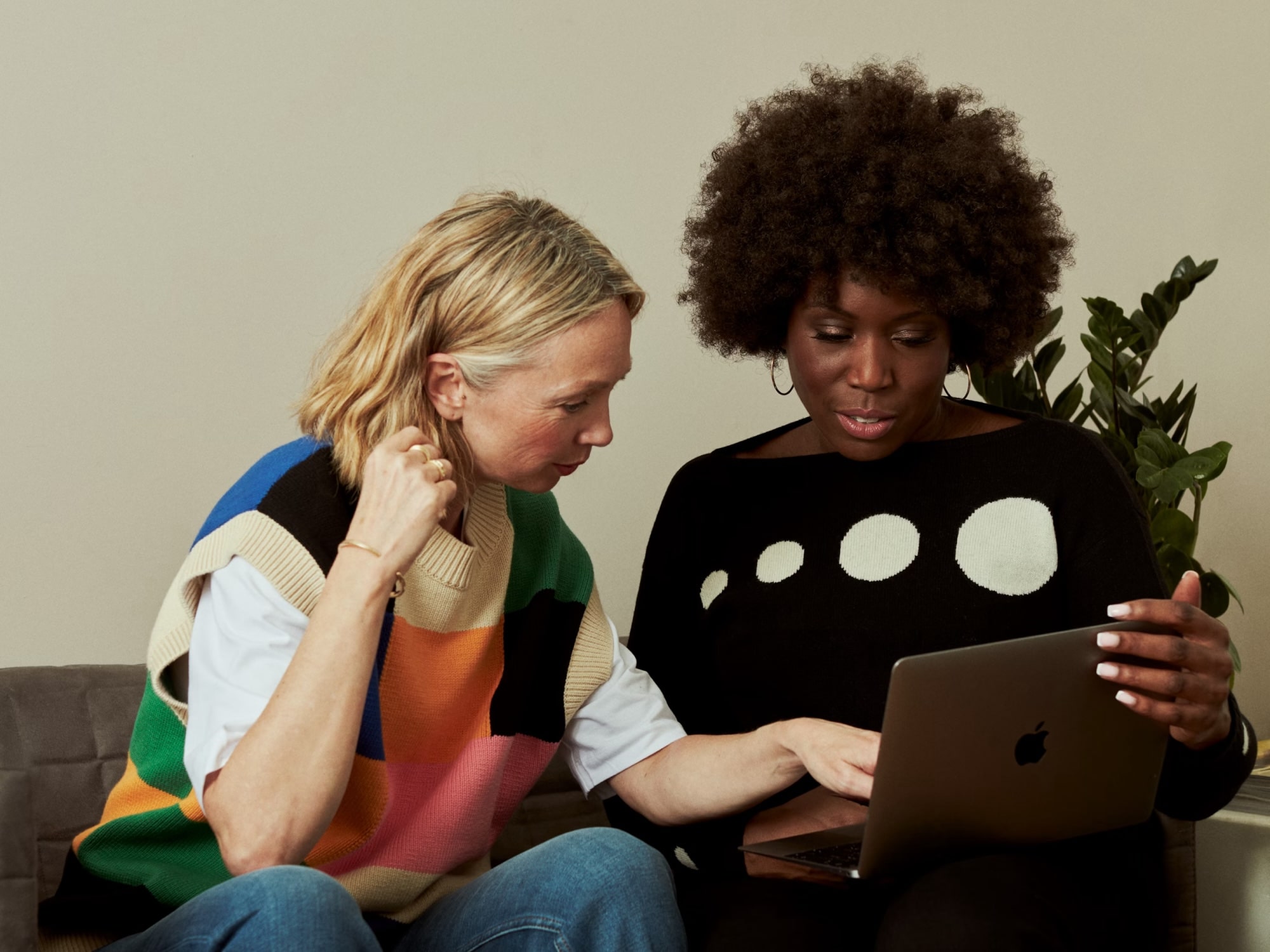 Two women sitting on a couch looking at a laptop, one wearing a colorful patchwork sweater vest and the other in a black sweater with white circles.