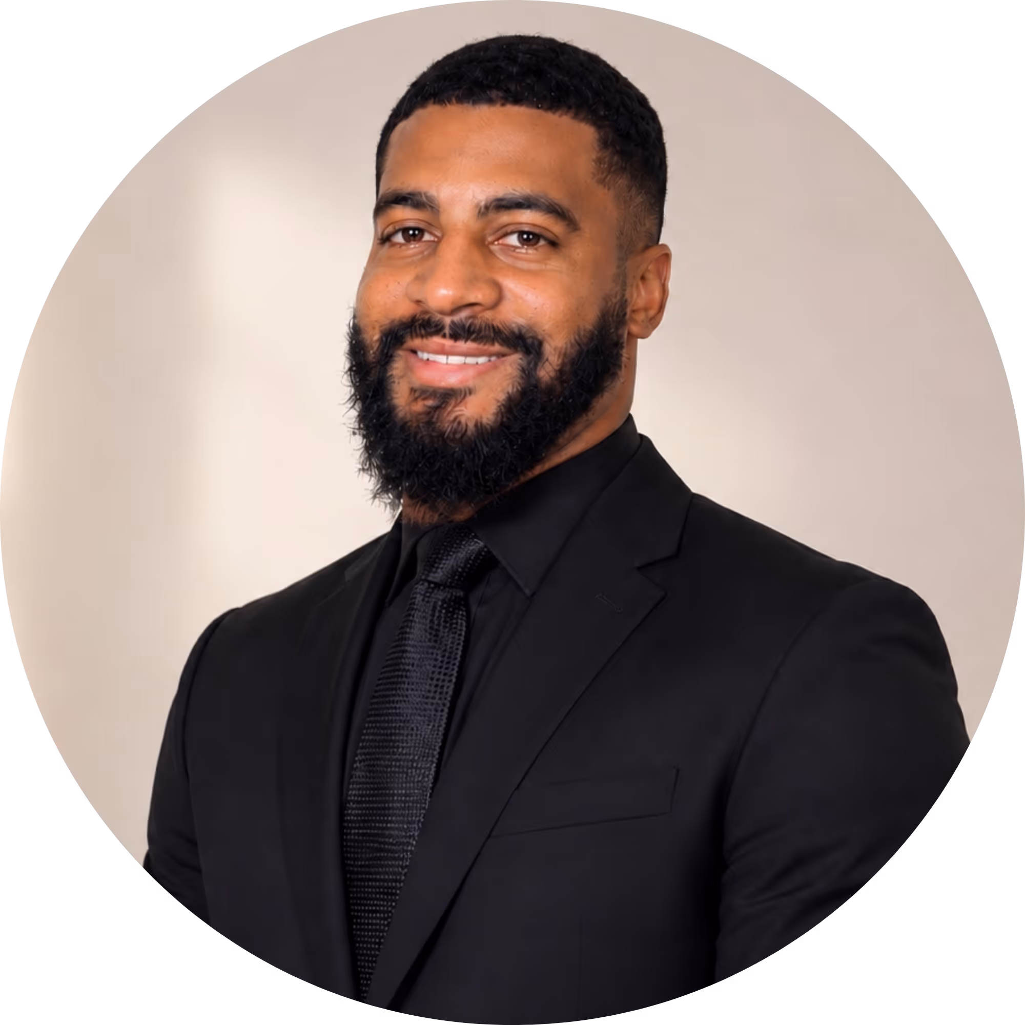 Smiling man with beard and dreadlocks wearing a navy blue suit jacket and white dress shirt against a plain light background.