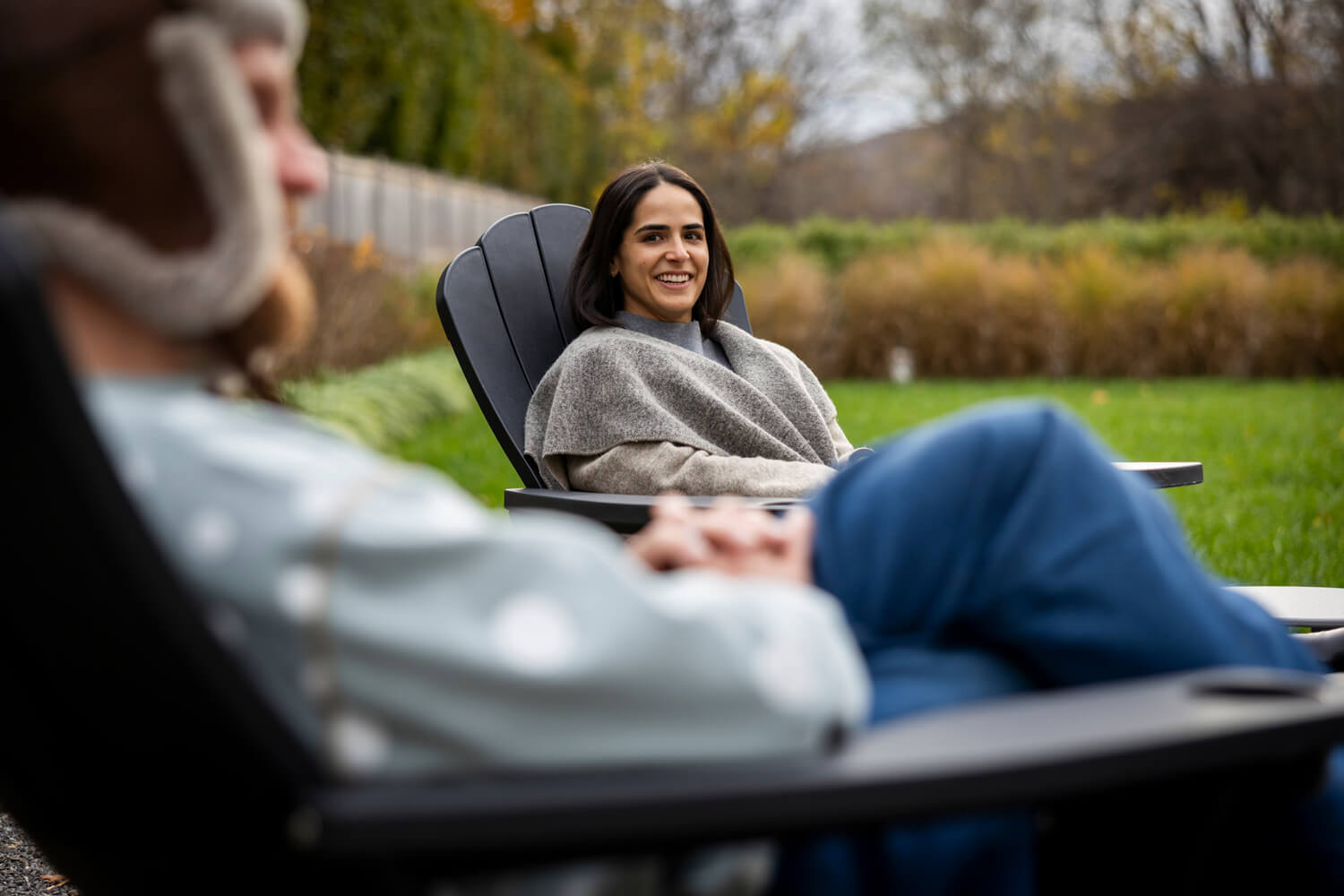 A woman wrapped in a grey shawl sits in a dark Adirondack chair, smiling toward someone out of focus in the foreground. They are outdoors on a green lawn with autumn trees and shrubs in the background.