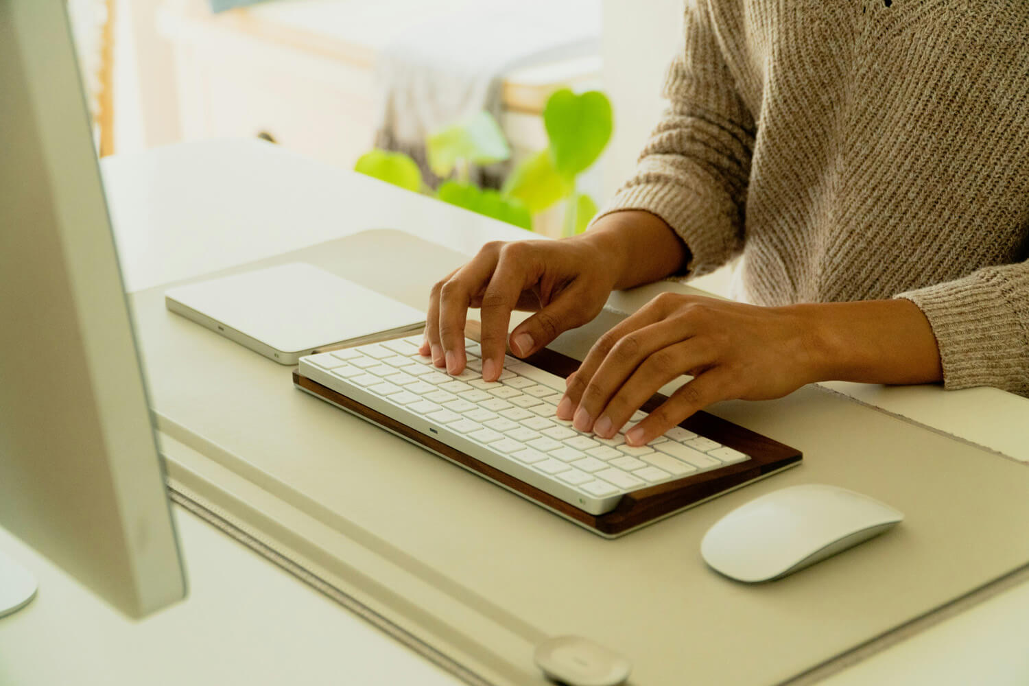 A close-up of a person's hands typing on a slim, white keyboard with a wooden base. The workspace is minimalist and bright, featuring a white desk, a matching computer monitor, a trackpad, and a mouse on a neutral-toned desk mat.