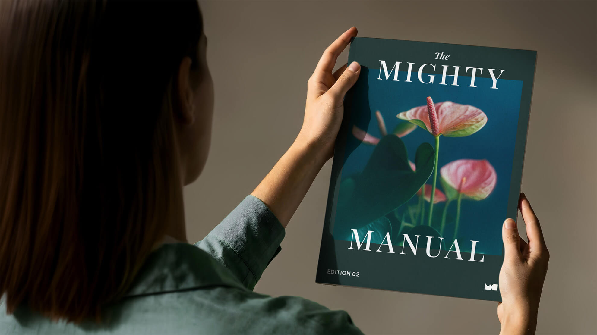 A woman seen from behind holds up a magazine titled "The Mighty Manual." The cover features a dark background with pink anthurium flowers. She is wearing a seafoam green shirt, and the scene is softly lit against a neutral wall.