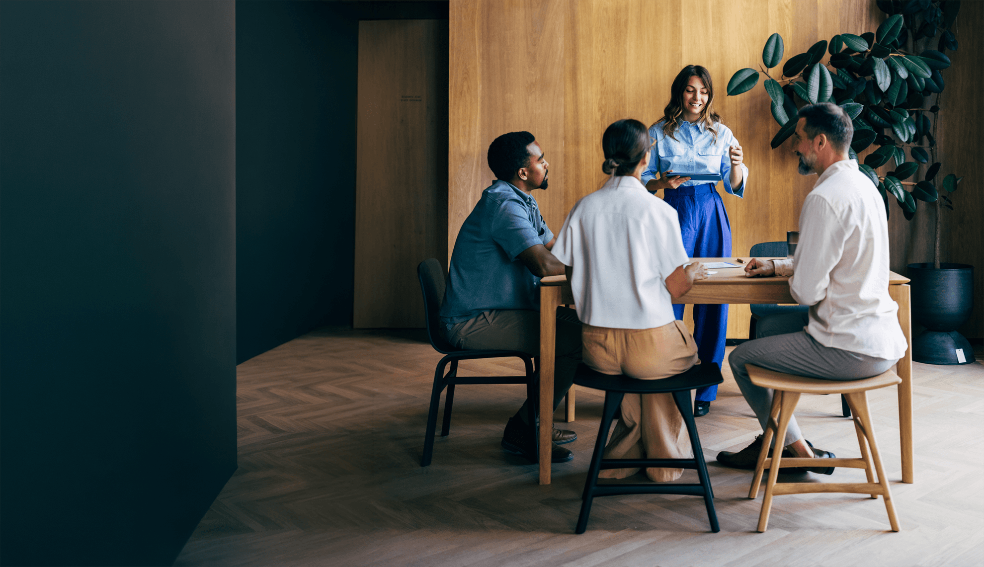 A woman standing at a table leads a meeting with three seated colleagues in a modern, wood-paneled office.