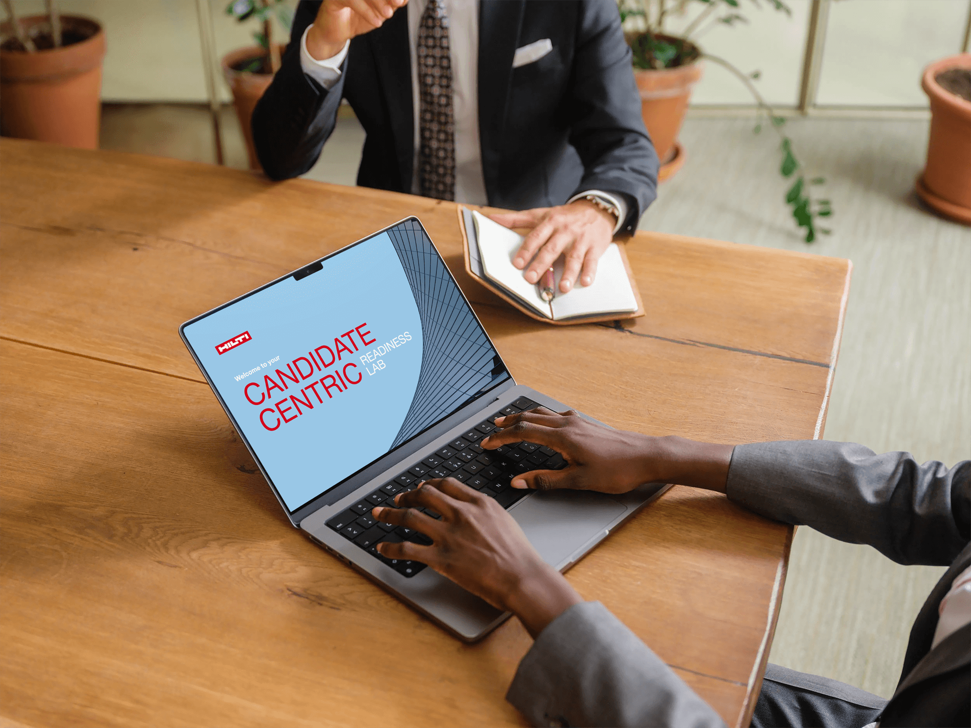 A person's hands typing on a laptop displaying a "Candidate Centric" presentation during a professional meeting at a wooden table.