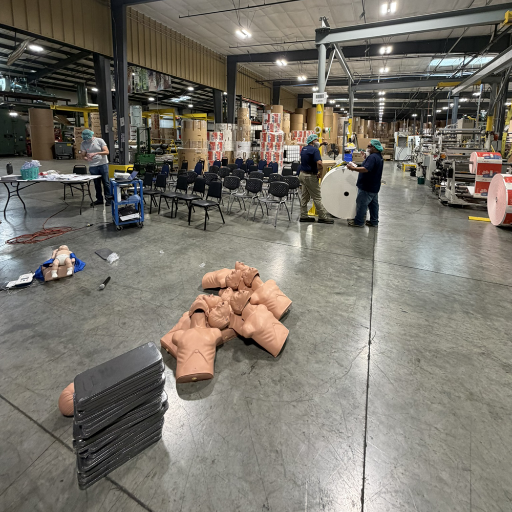 CPR manikins, training mats and an AED trainer staged on a manufacturing floor for an onsite First Aid and CPR class.