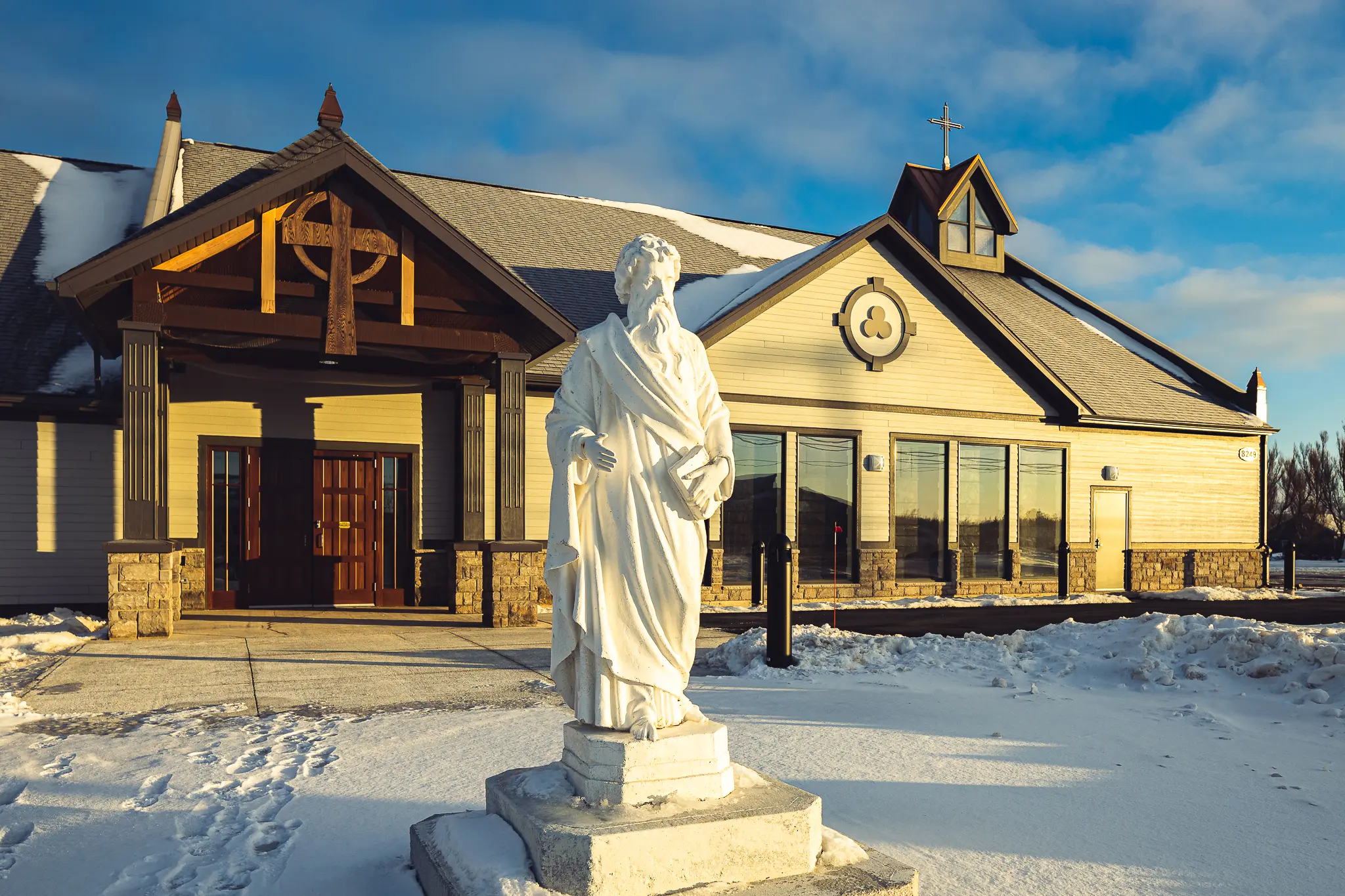 A white statue of a religious figure stands in front of a church with snow-covered ground and a clear blue sky.