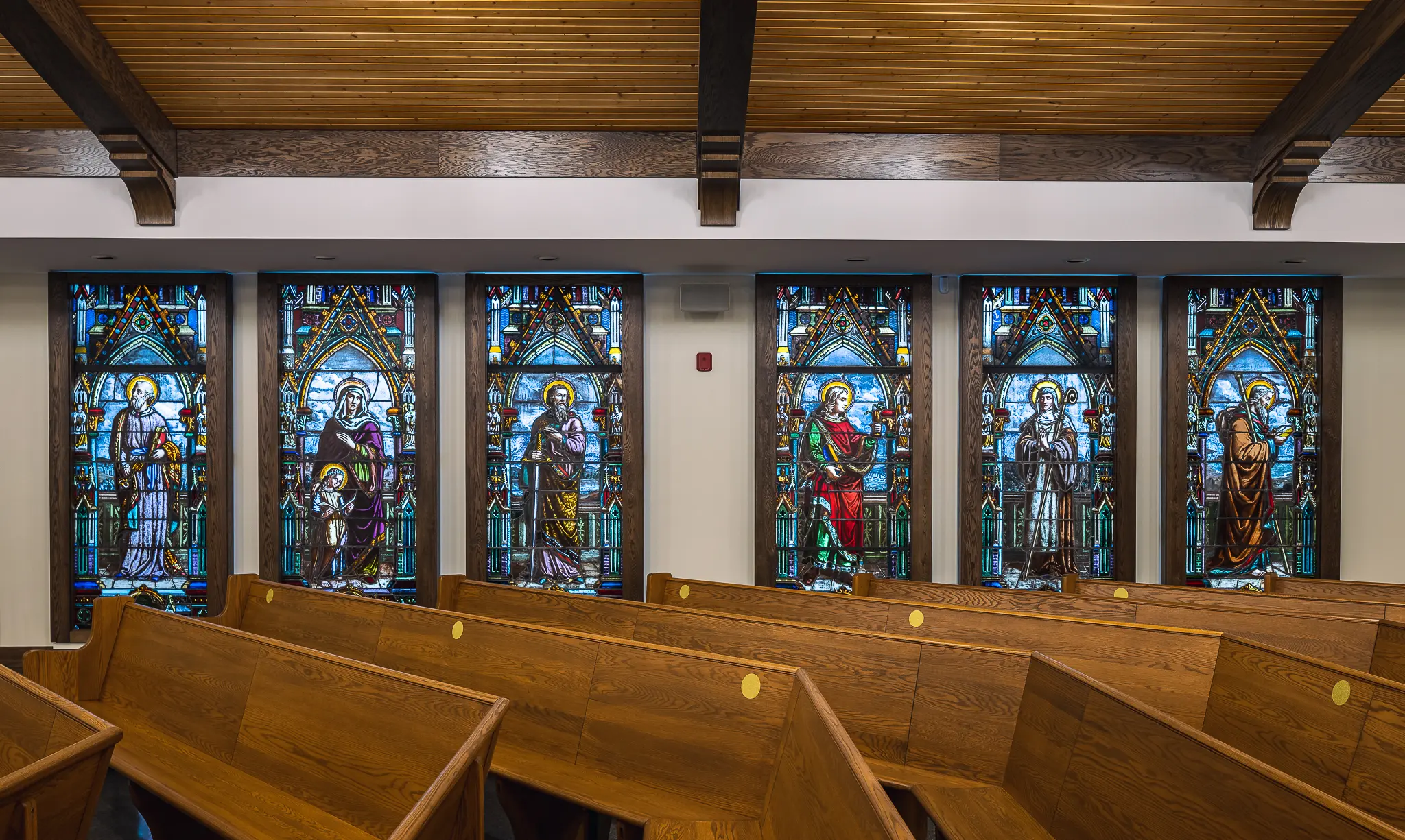Stained glass windows depicting saints in a church interior with wooden pews and a vaulted wooden ceiling.