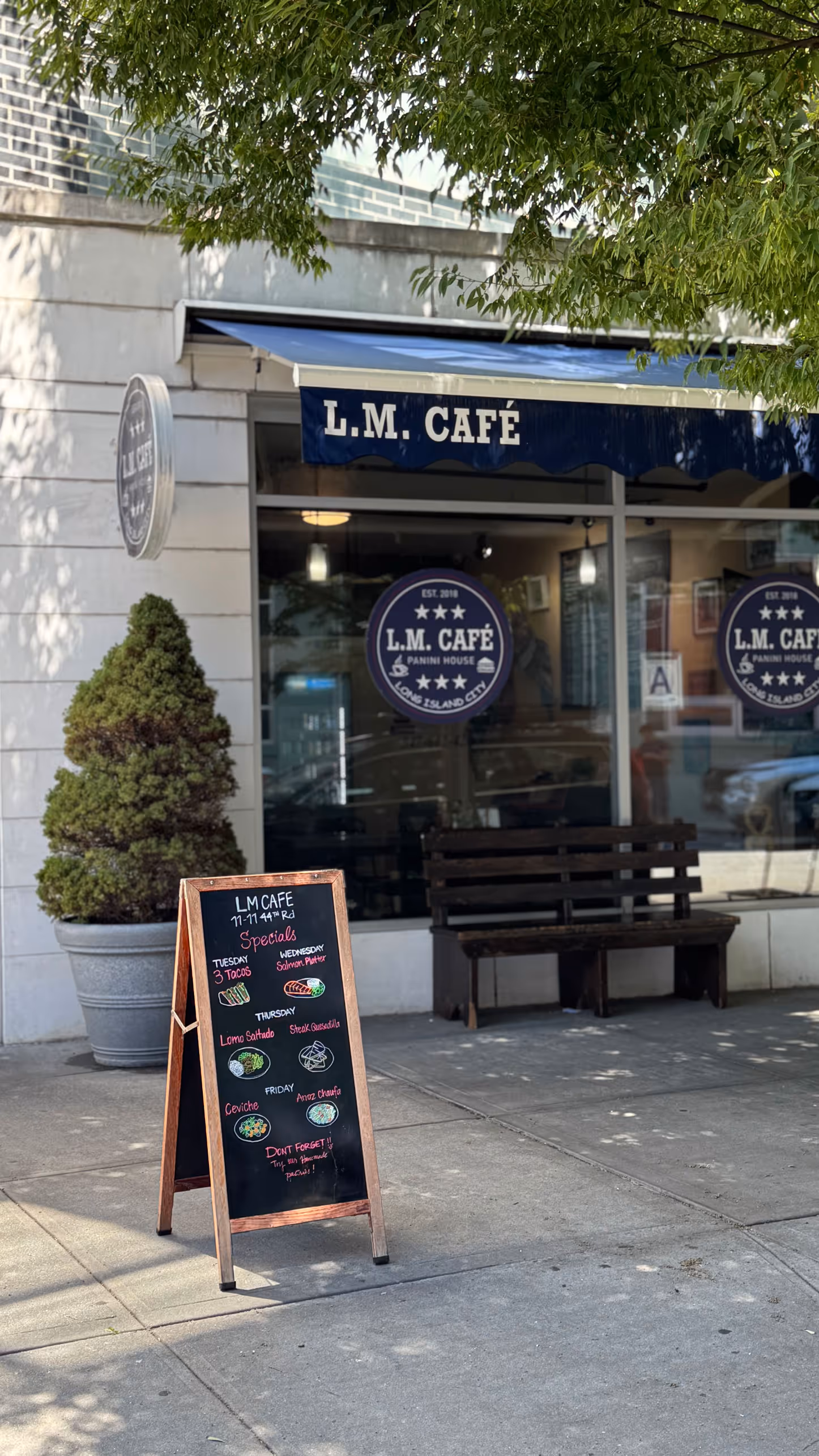 Sidewalk view of L.M. Café with a blue awning, a wooden bench, a large potted plant, and a chalkboard displaying weekly food specials.