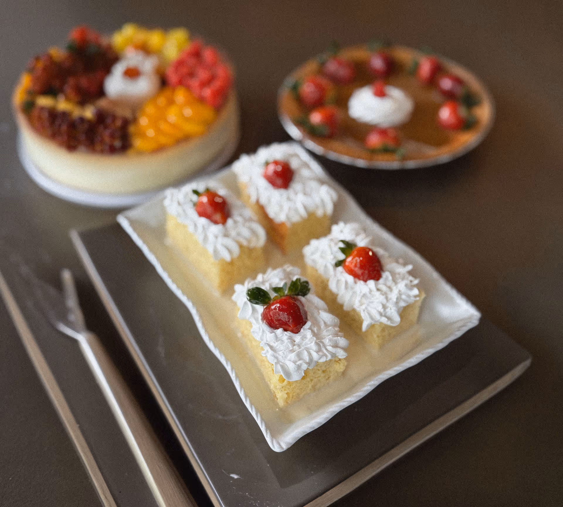 Four pieces of sponge cake topped with whipped cream and a strawberry on a rectangular white plate, with a fruit cake and a cherry-topped pie in the background.
