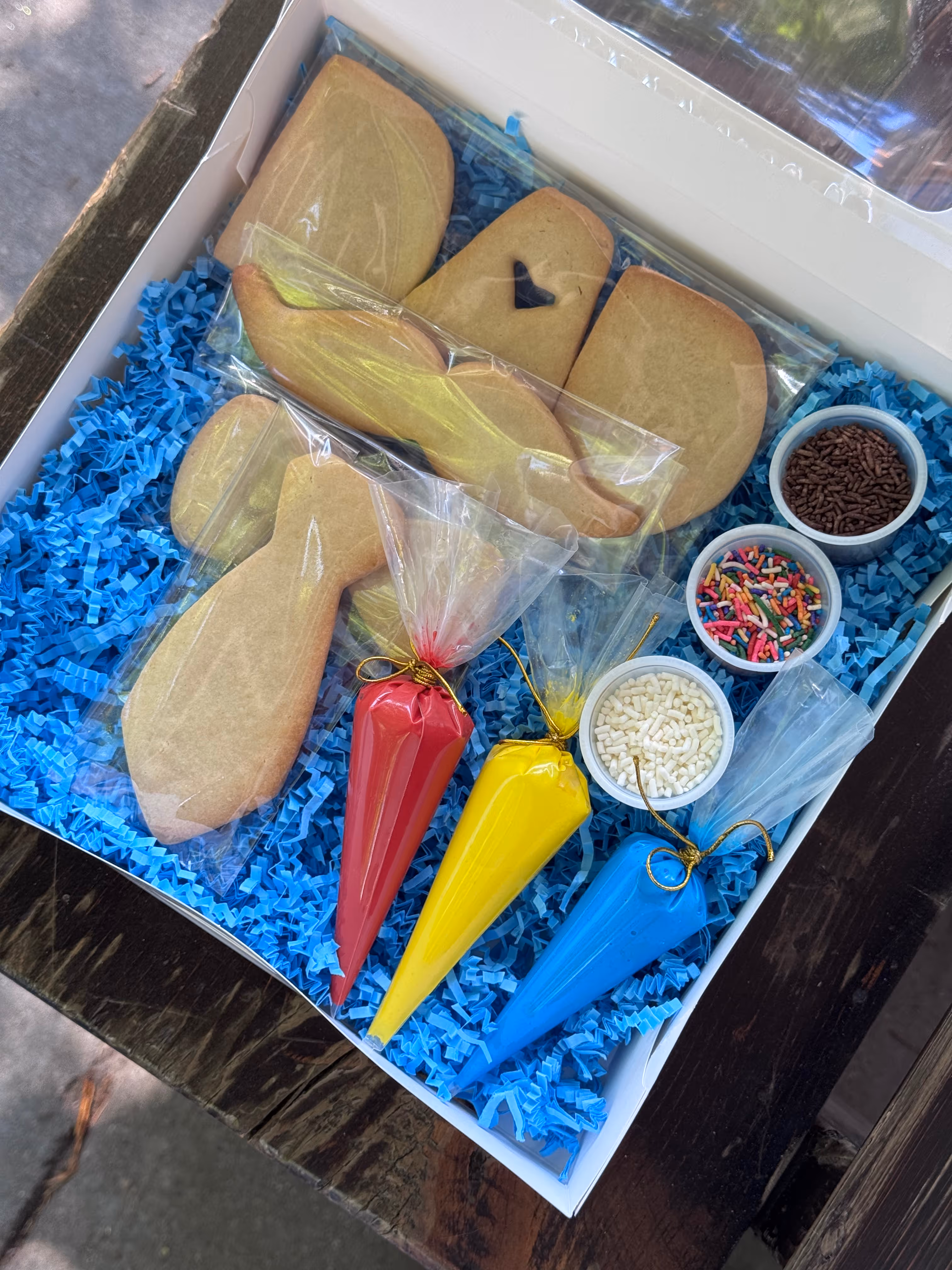 Box with uniced cookies, red, yellow, and blue icing bags, and containers of multicolored sprinkles.