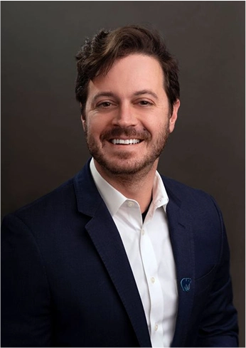 Smiling man with dark hair and beard wearing a navy blazer and white shirt against a dark background.