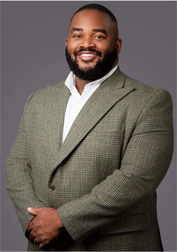 Smiling man with beard in a green checked blazer and white shirt standing against a gray background.