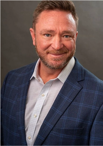 Smiling middle-aged man with short brown hair and beard wearing a blue checkered blazer and white shirt against a plain gray background.