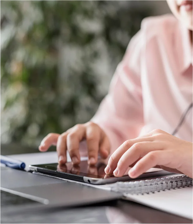Person typing on a tablet placed on a notebook beside a laptop on a desk.