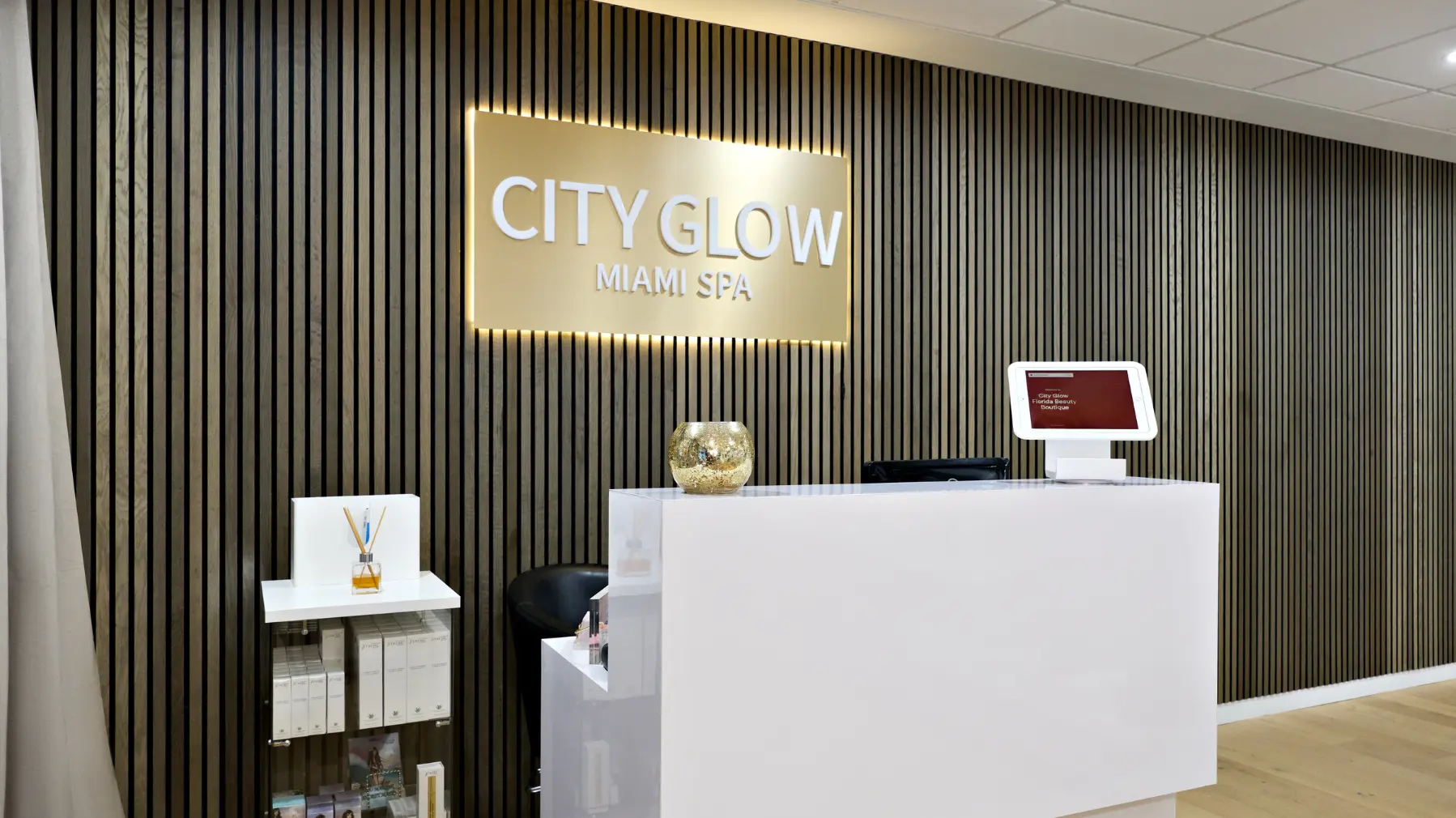 A clean view of the modern reception desk at City Glow Beauty Salon in Brickell, Miami. It features a textured wood-slat wall with a large backlit gold sign that reads 'CITY GLOW MIAMI SPA.' A white desk, reed diffuser, and boxed skincare products are visible, creating a welcoming atmosphere.