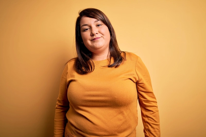 Smiling woman with dark brown hair wearing a mustard yellow long-sleeve shirt standing against a matching yellow background.