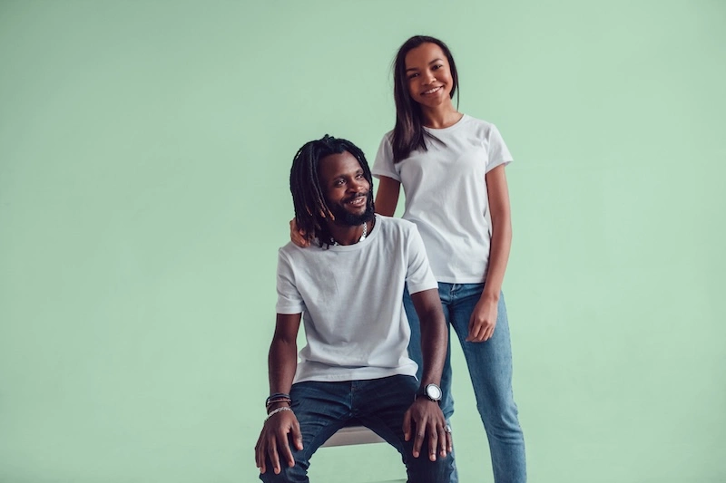 Smiling couple wearing white t-shirts and jeans, with the woman standing behind the seated man against a light green background.