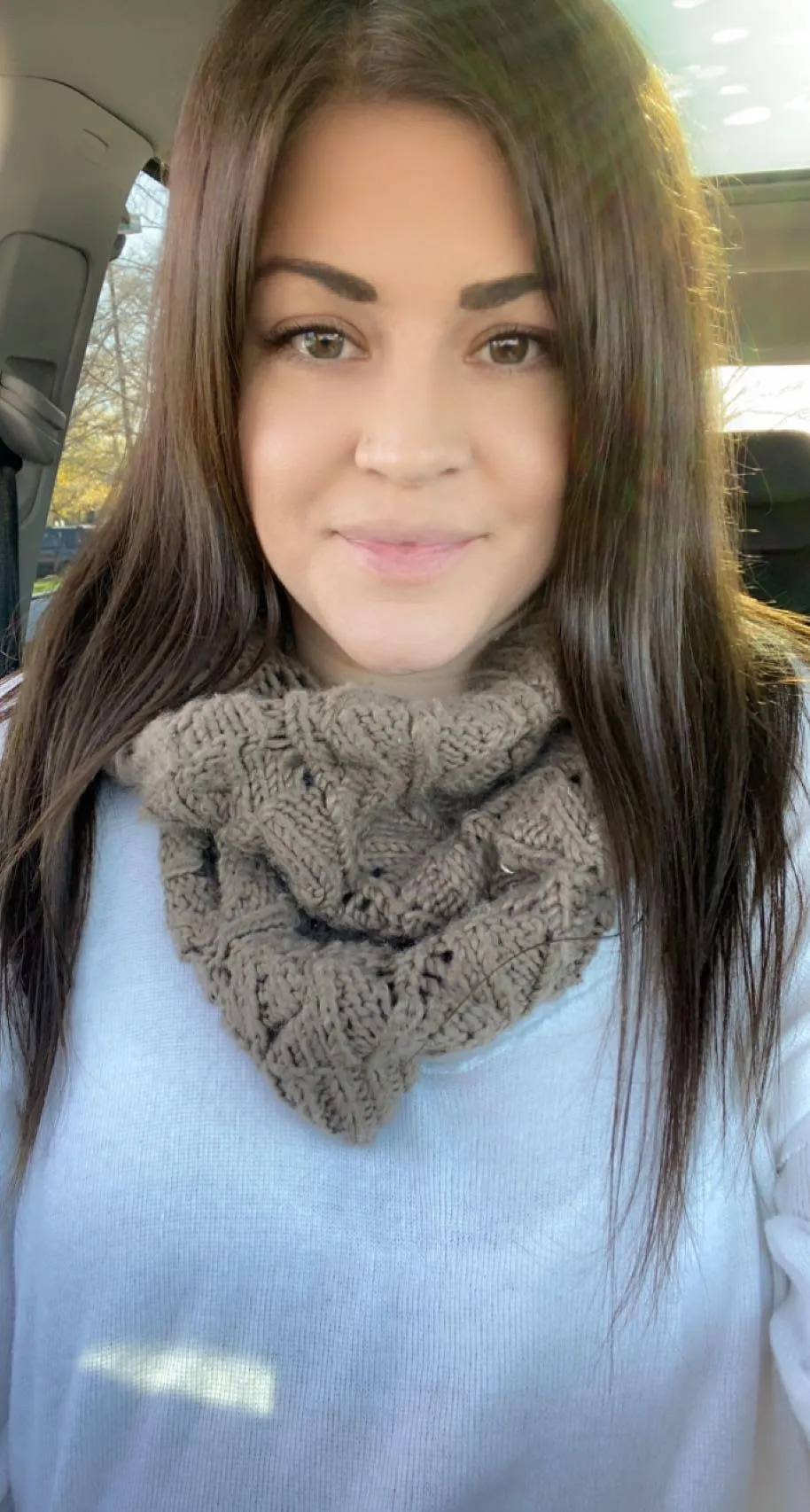 Woman with long brown hair wearing a beige knitted scarf and a white sweater, sitting inside a car.
