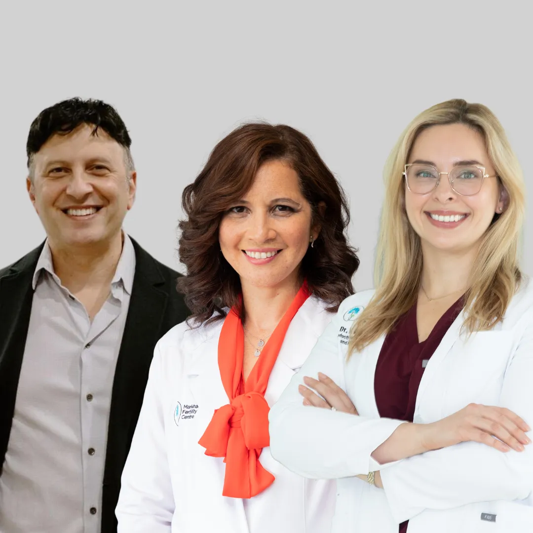 Three smiling medical professionals standing side by side against a gray background, two women in white lab coats and one man in a dark blazer.