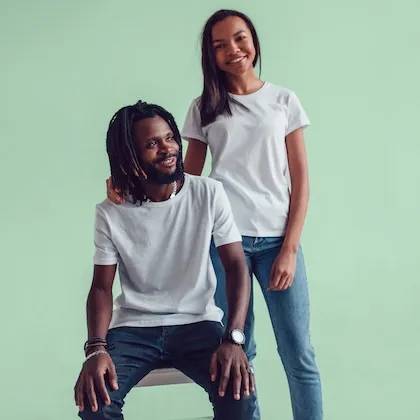 Smiling couple wearing white t-shirts and jeans, with the woman standing behind the seated man against a light green background.
