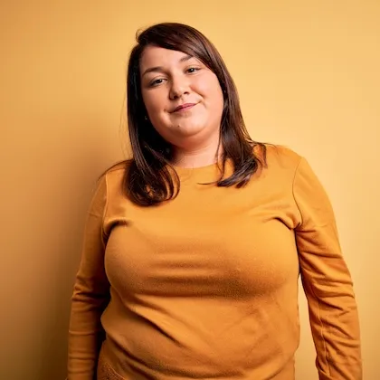 Smiling woman with dark brown hair wearing a mustard yellow long-sleeve shirt standing against a matching yellow background.